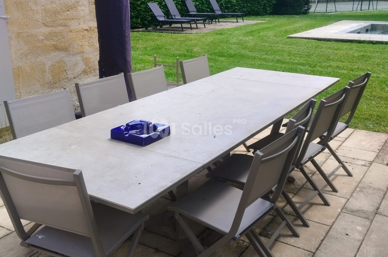 Table de jardin rectangulaire entourée de chaises pliantes grises, située sur une terrasse en pierre près d'une pelouse.