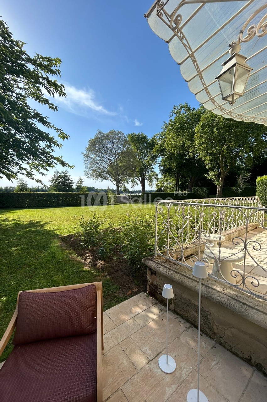 Vue d'une terrasse ensoleillée avec fauteuil, lampes et jardin verdoyant bordé d'arbres sous un ciel bleu.