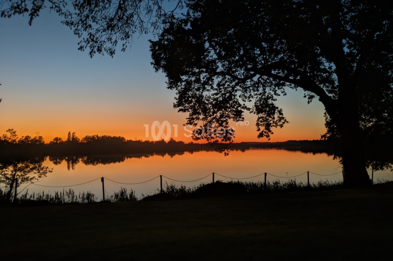 Paysage au crépuscule avec un lac calme reflétant un ciel orangé, entouré de silhouettes d'arbres et d'une clôture.