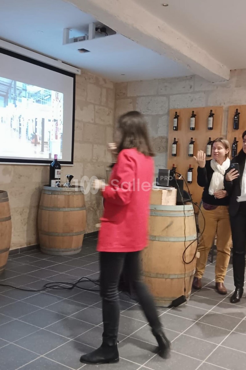 Une femme en rouge marche vers un écran de projection dans une salle avec des tonneaux et des bouteilles de vin exposées.