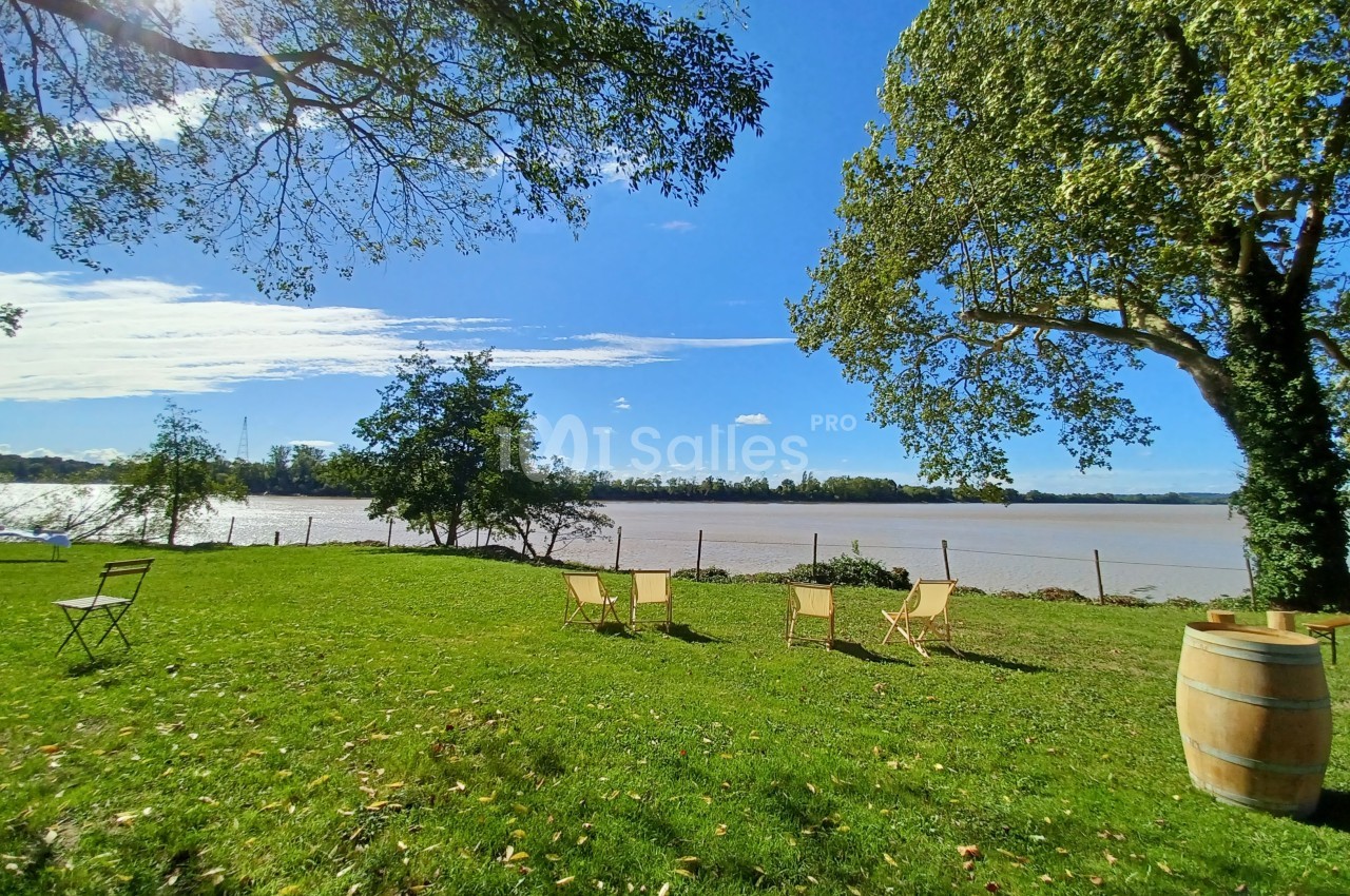 Chaises et table disposées sur une pelouse verdoyante près d'un fleuve, entourées d'arbres sous un ciel bleu.