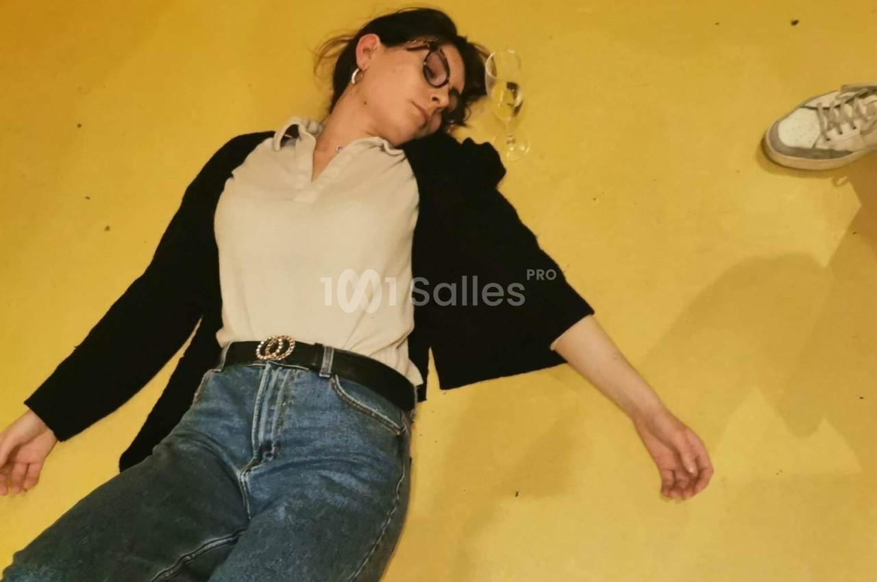 Femme allongée sur le sol jaune, lunettes tombées à côté d'elle, avec une chaussure visible en arrière-plan.