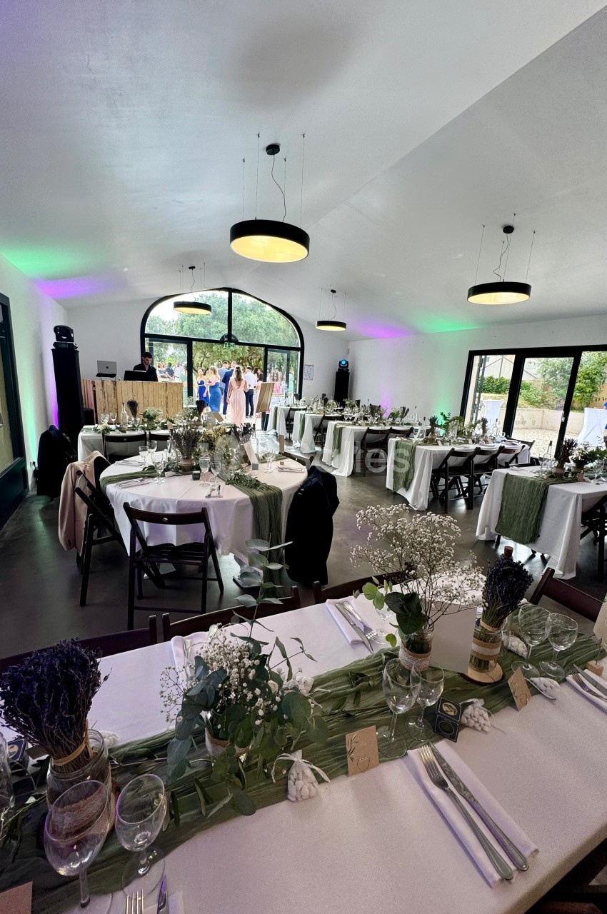 Salle de réception décorée avec des tables dressées, ornées de fleurs et de nappes blanches, éclairée par des suspensions.
