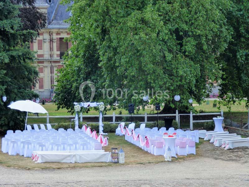 Chaises blanches disposées en extérieur pour une cérémonie, sous des arbres avec des décorations et une arche fleurie.