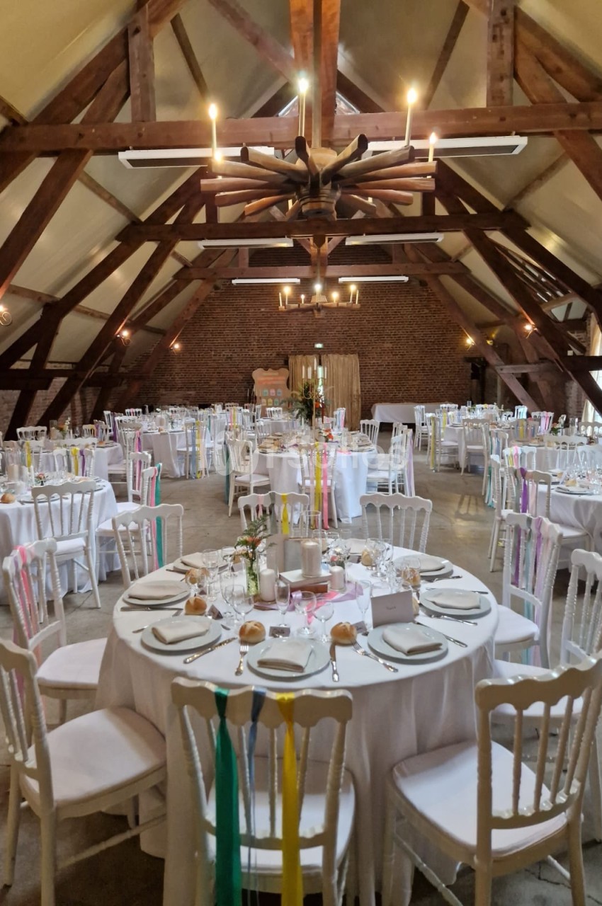Salle de réception avec tables rondes dressées, décorées de rubans colorés et éclairée par des lustres en bois.
