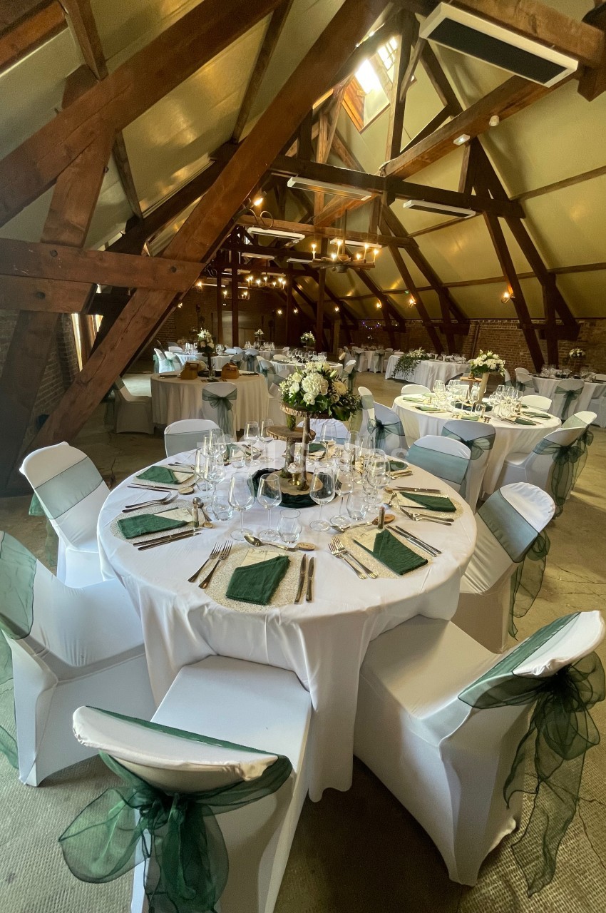 Salle de réception avec poutres apparentes, tables rondes dressées avec nappes blanches et décorations florales.