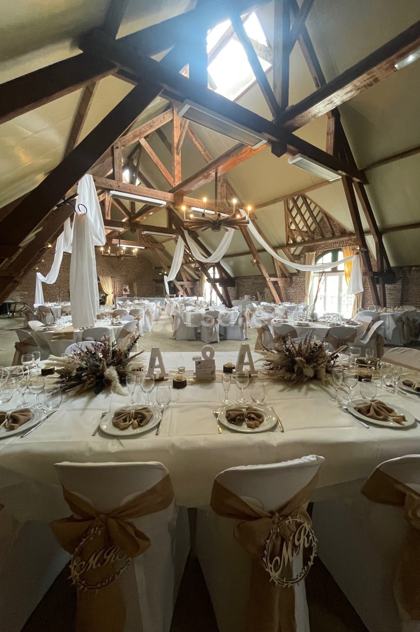 Salle de réception décorée avec des tables dressées, nappes blanches, chaises ornées de rubans et poutres apparentes.