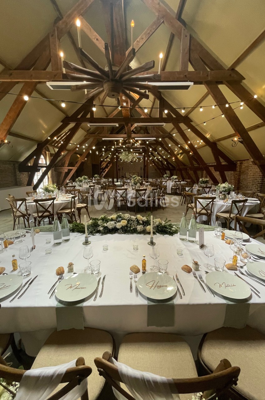 Salle de réception décorée pour un mariage, avec tables dressées, chaises en bois et guirlandes lumineuses suspendues.