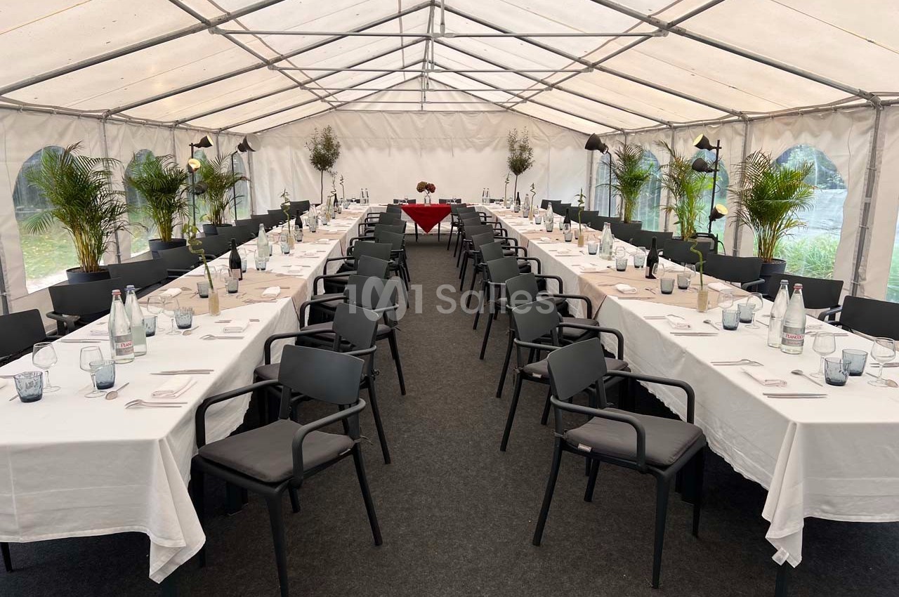 Tables dressées avec nappes blanches et chaises noires alignées dans une grande tente, décorée de plantes vertes.