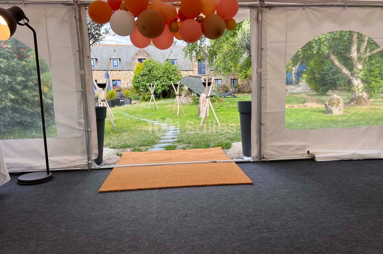 Vue depuis l'intérieur d'une tente avec des ballons suspendus, donnant sur un jardin et des bâtiments en pierre.
