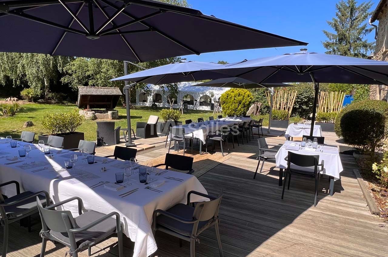 Terrasse ensoleillée avec tables dressées sous des parasols, entourée de verdure et d'une pelouse arborée.