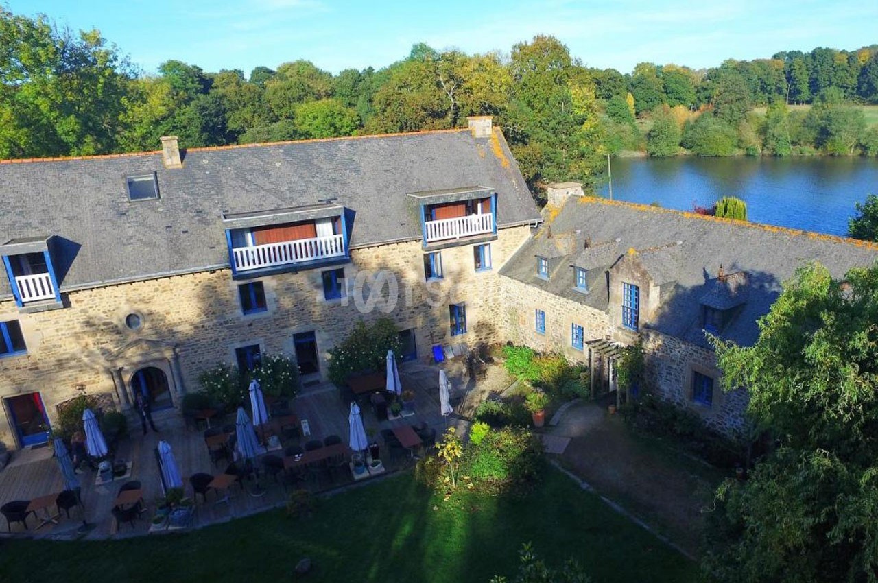 Vue aérienne d'un bâtiment en pierre avec terrasse et tables extérieures, entouré de verdure près d'un plan d'eau.