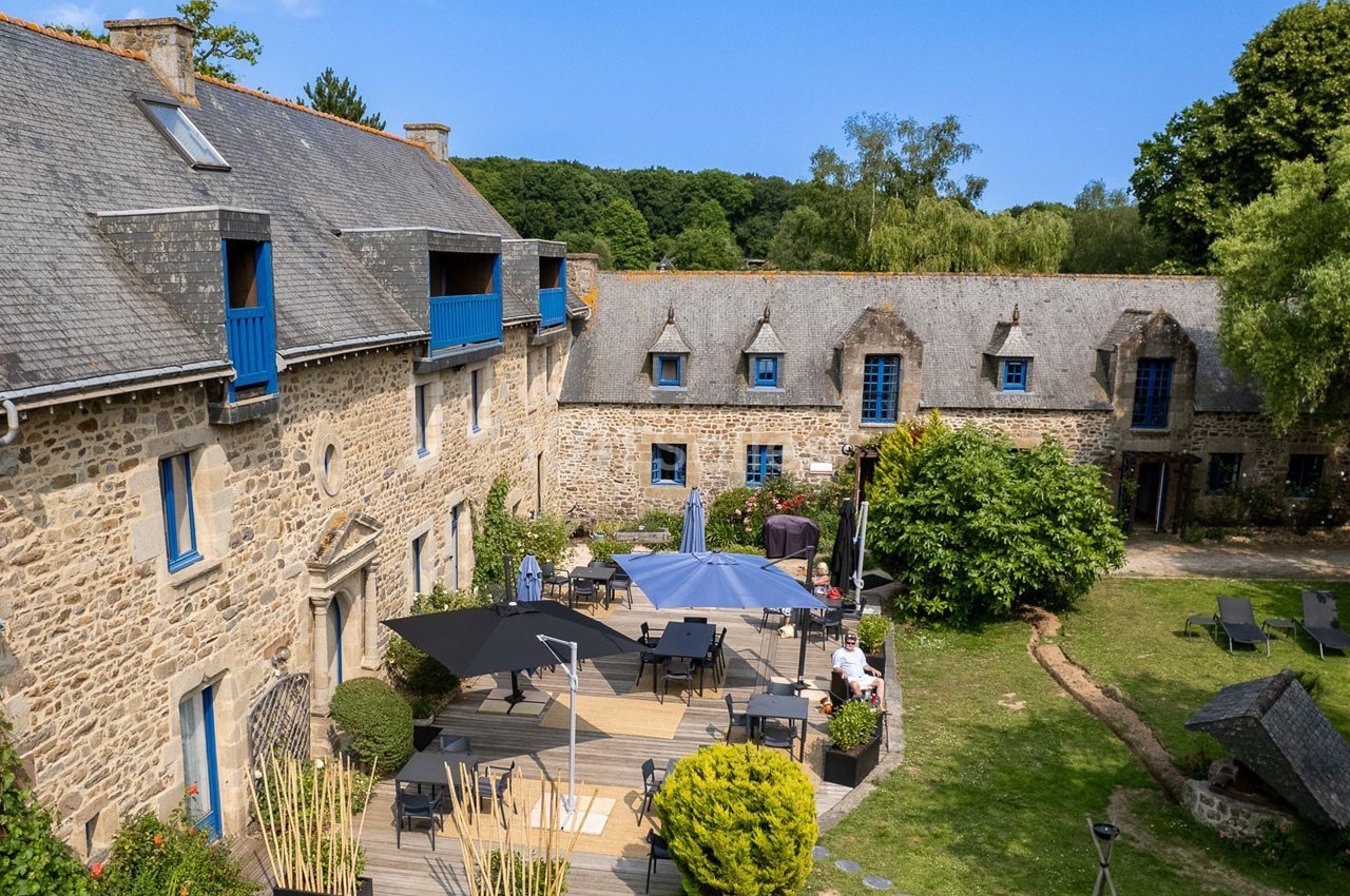 Cour intérieure d'un bâtiment en pierre avec terrasse aménagée, parasols, chaises longues et végétation environnante.