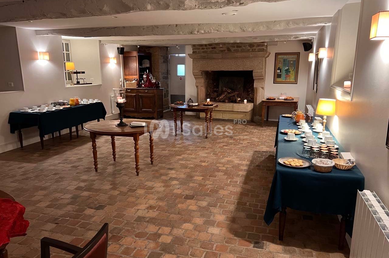 Salle avec tables dressées pour un buffet, cheminée en pierre au fond, éclairage chaleureux et sol en carreaux.