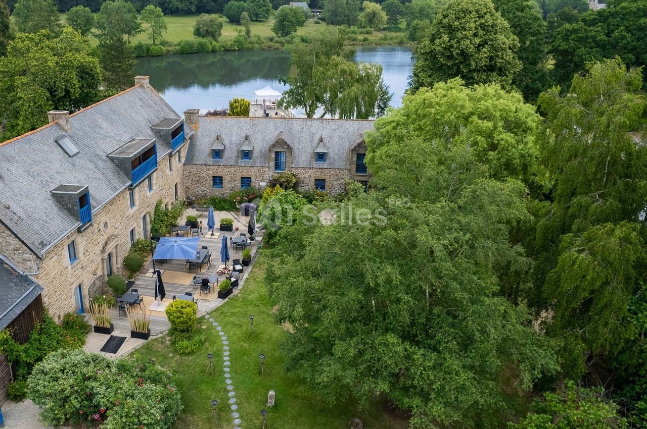 Vue aérienne d'un bâtiment en pierre entouré de verdure, avec une terrasse aménagée près d'un plan d'eau.
