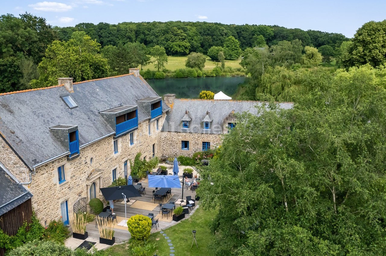 Vue aérienne d'un bâtiment en pierre avec terrasse aménagée, entouré de verdure et d'un plan d'eau en arrière-plan.