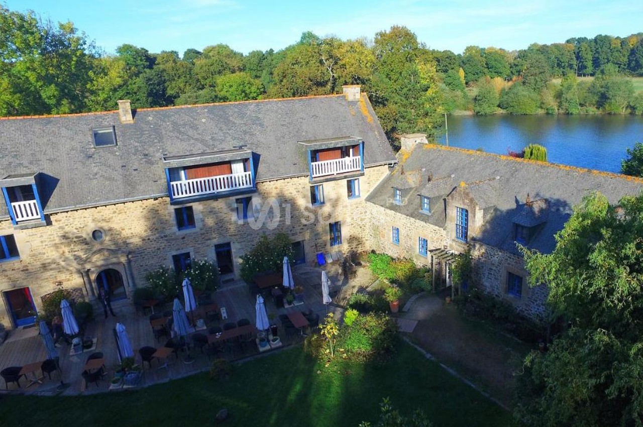 Vue aérienne d'un bâtiment en pierre avec terrasse et tables extérieures, entouré de verdure près d'un plan d'eau.