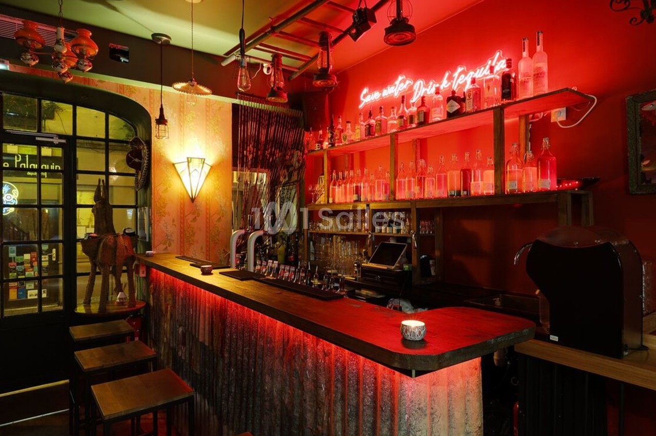 Bar à l'ambiance chaleureuse avec éclairage rouge, comptoir en bois, étagères de bouteilles et néon lumineux au mur.