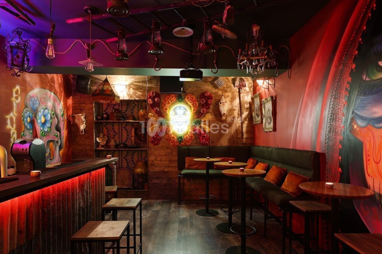 Bar à l’ambiance tamisée avec des décorations murales colorées, des tables hautes et un éclairage rouge chaleureux.