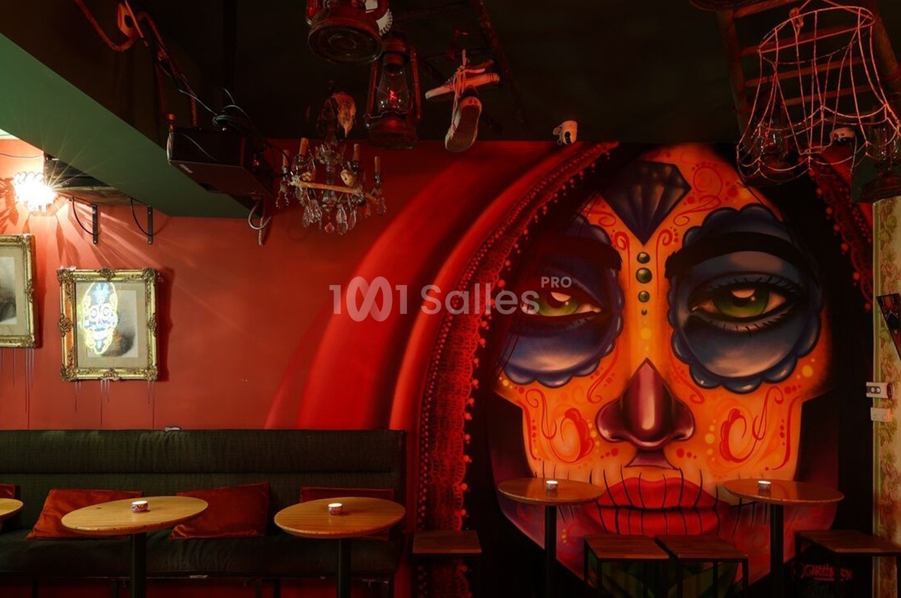 Mur décoré d'une fresque colorée représentant un visage maquillé, entouré de tables et de cadres anciens.