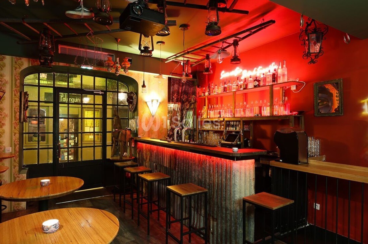 Intérieur d'un bar avec comptoir en métal, tabourets hauts, éclairage néon rouge et décorations murales éclectiques.