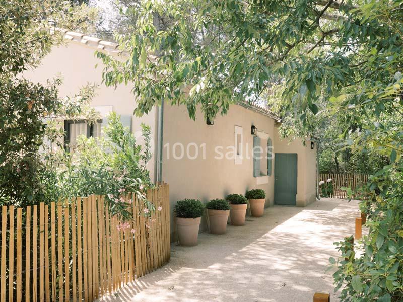 Maison en pierre claire entourée de végétation, avec une clôture en bois et des pots de fleurs alignés devant l'entrée.