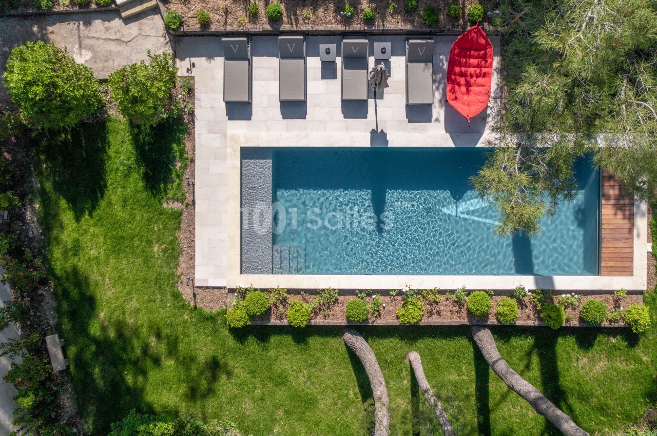 Vue aérienne d'une piscine rectangulaire entourée de chaises longues, d'un parasol rouge et de verdure.