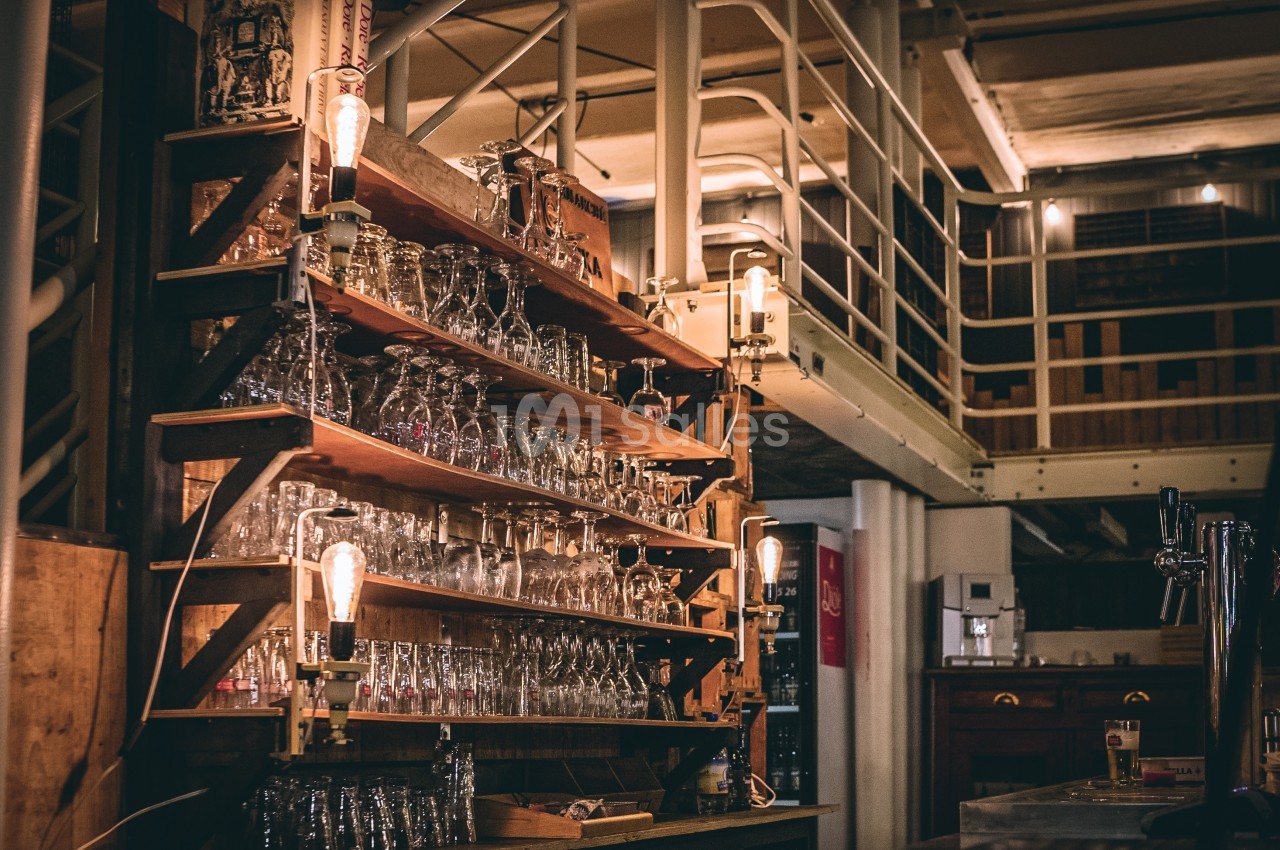 Étagères en bois remplies de verres dans un bar à l'ambiance industrielle avec éclairage tamisé.