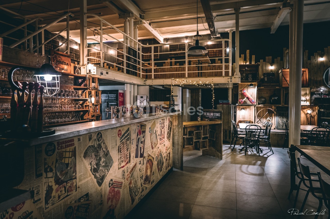 Intérieur d'un bar au style industriel avec un comptoir décoré de journaux et un espace aménagé en mezzanine.