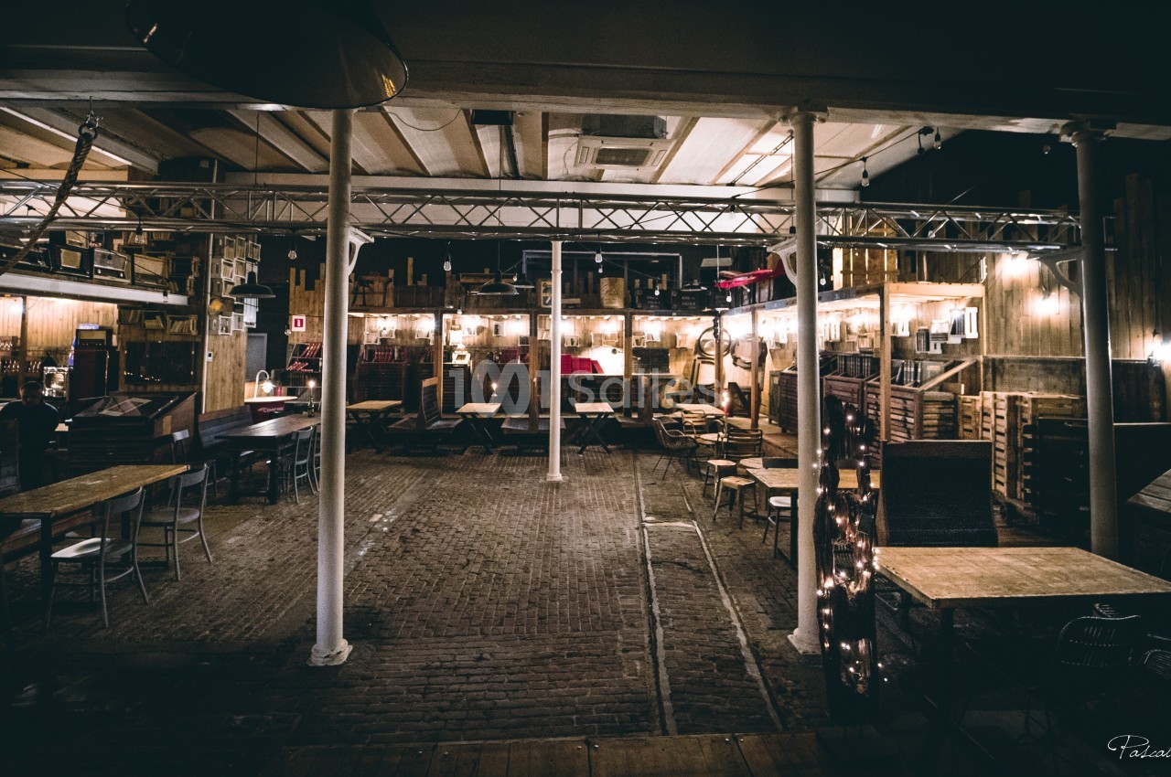Salle intérieure avec tables et chaises en bois, éclairée par des lumières chaleureuses, ambiance rustique et industrielle.