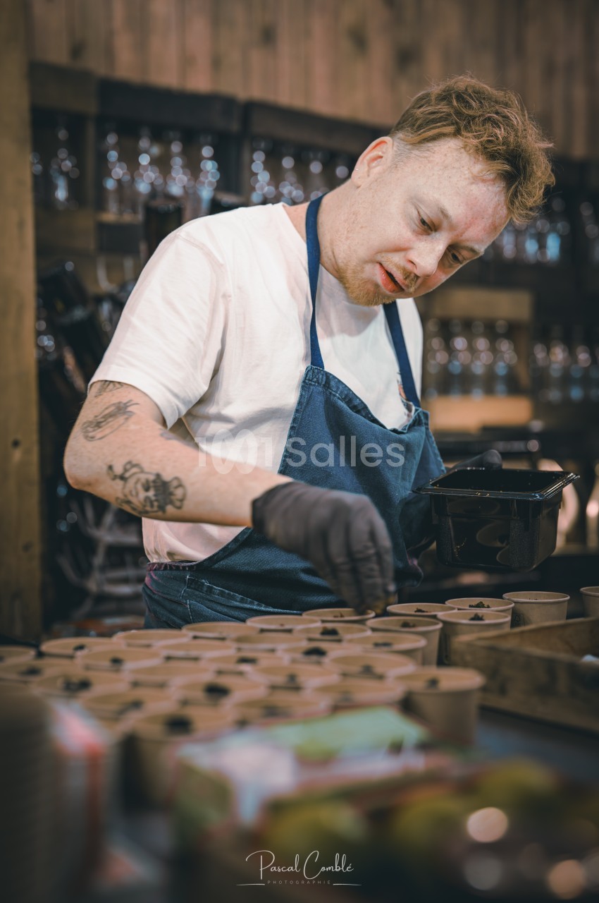 Homme en tablier bleu préparant des plats dans des contenants individuels sur un plan de travail en bois.