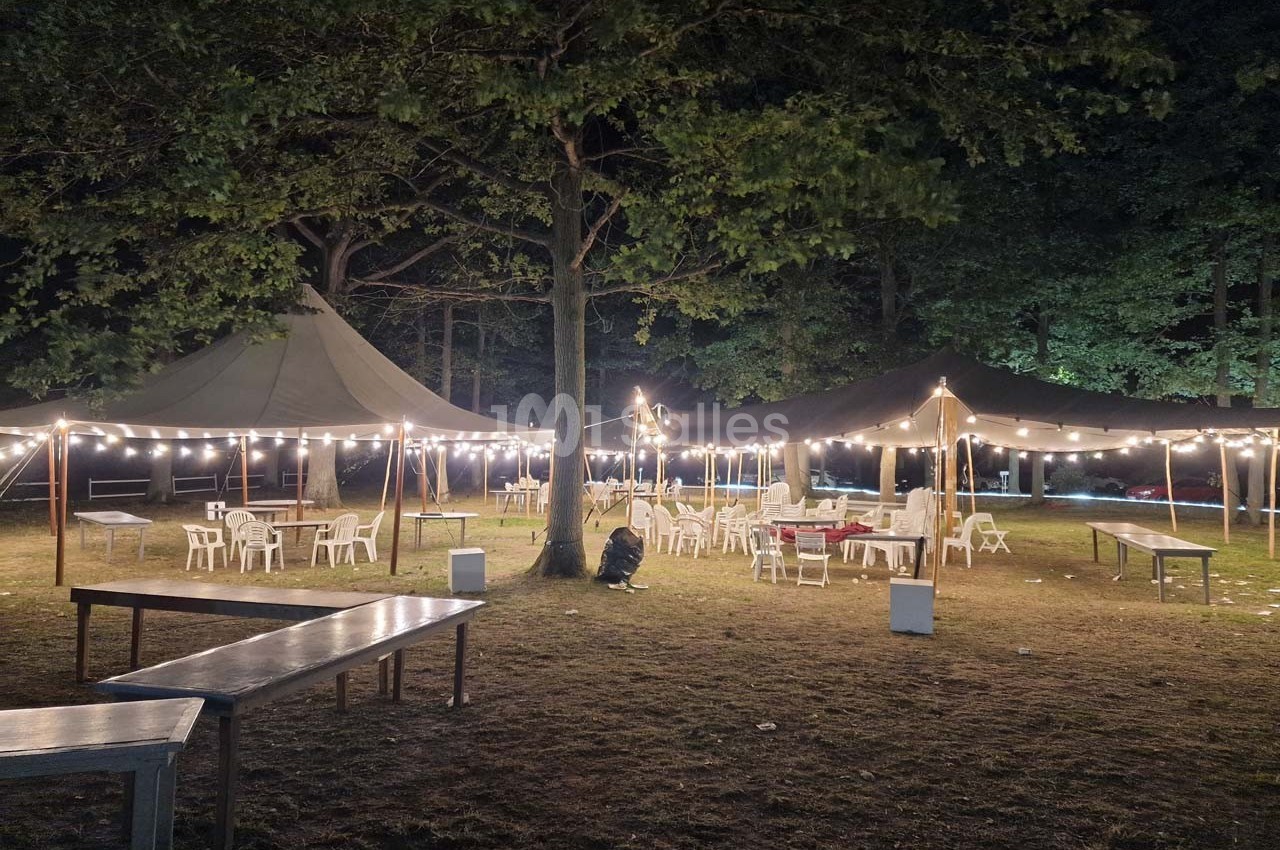 Tentes éclairées par des guirlandes lumineuses dans un espace extérieur avec des tables et des chaises disposées sur l'herbe.