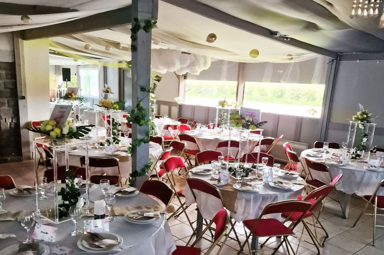 Salle décorée pour un événement avec des tables rondes dressées, nappes blanches, chaises rouges et ornements floraux.