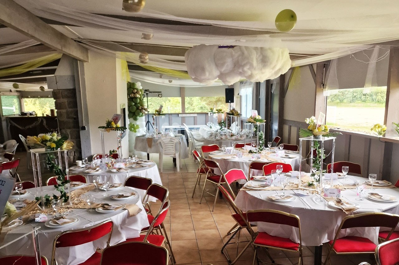 Salle décorée pour un événement avec des tables rondes dressées, chaises rouges et décorations suspendues.