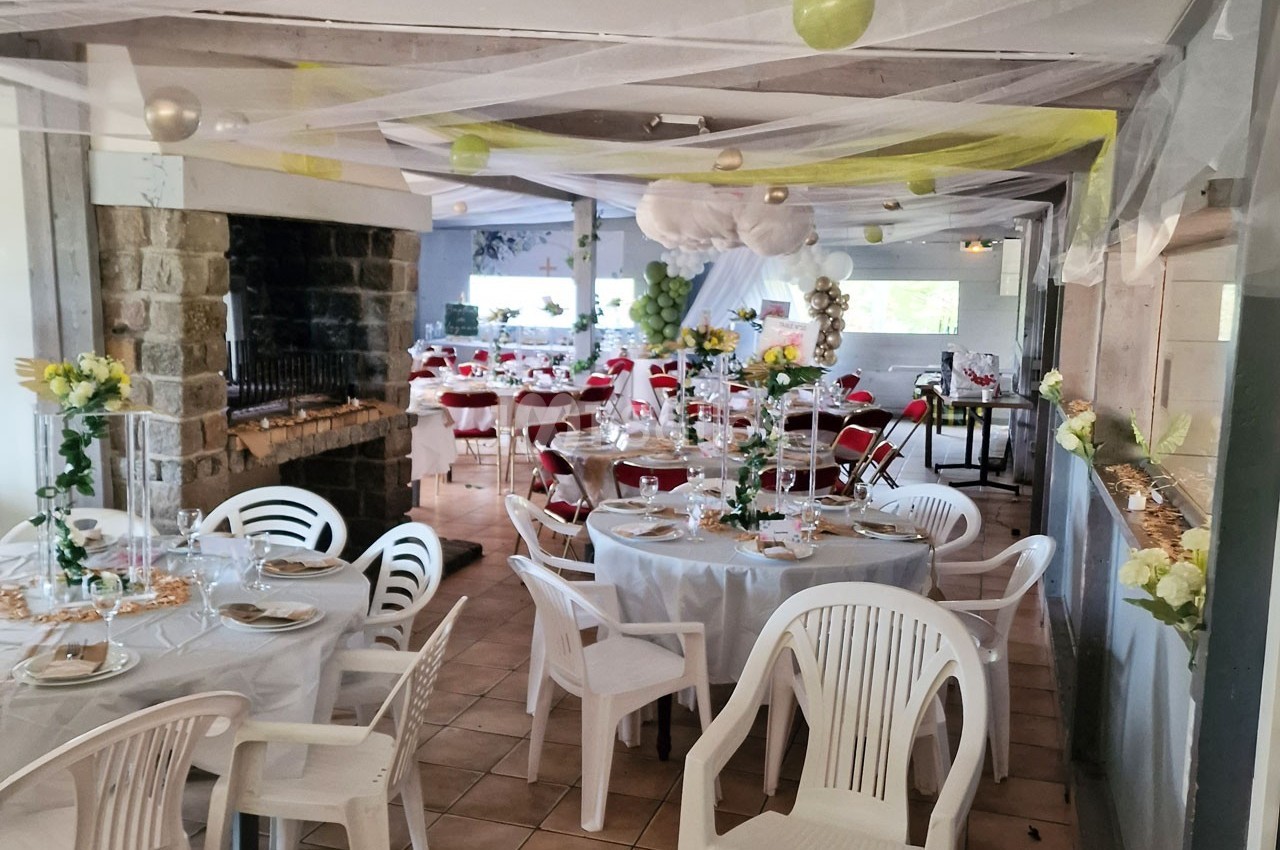 Salle décorée pour un événement avec tables rondes dressées, chaises blanches et ornements suspendus.