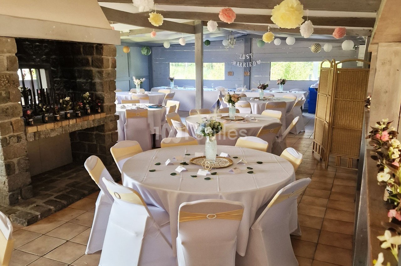 Salle décorée pour une réception avec tables rondes dressées, chaises blanches et ornements suspendus.