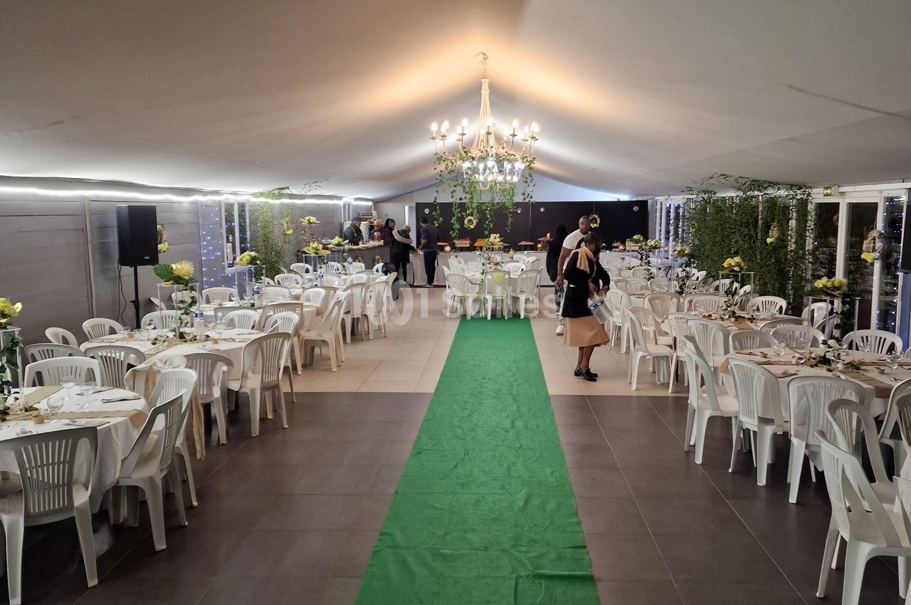Salle de réception décorée avec des tables rondes, des chaises blanches, un tapis vert et un lustre central.