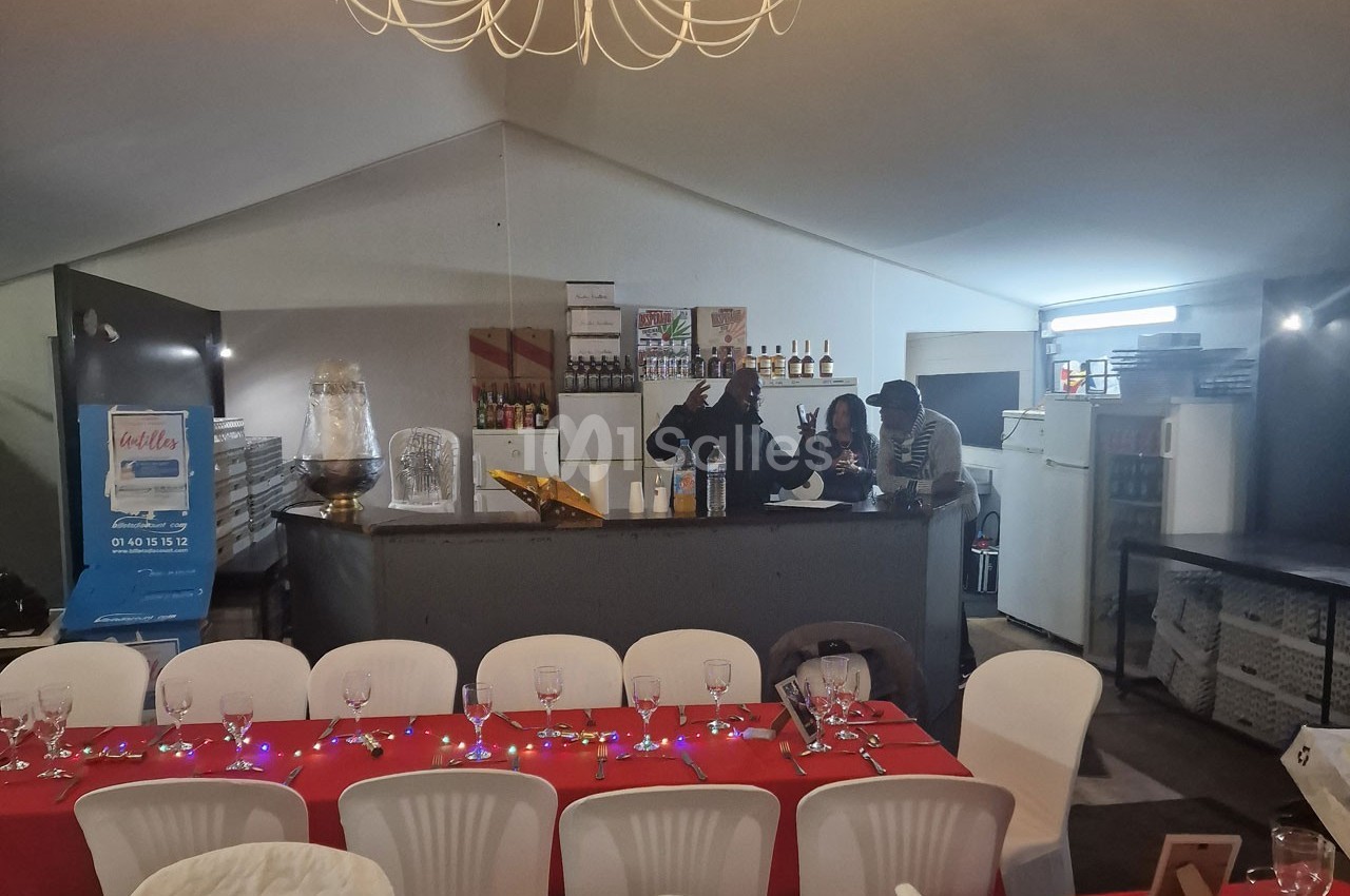 Salle de réception avec tables dressées en rouge et blanc, bar en arrière-plan avec trois personnes discutant.