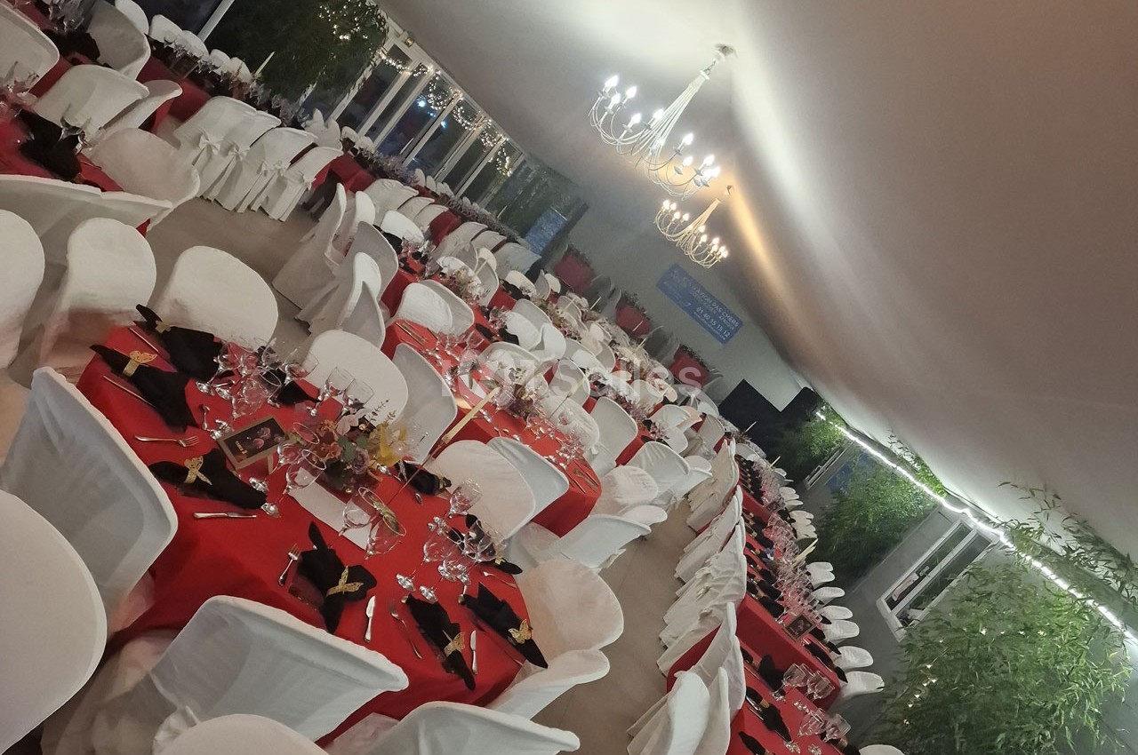 Salle de réception décorée avec des tables rondes couvertes de nappes rouges, chaises blanches et lustres suspendus.