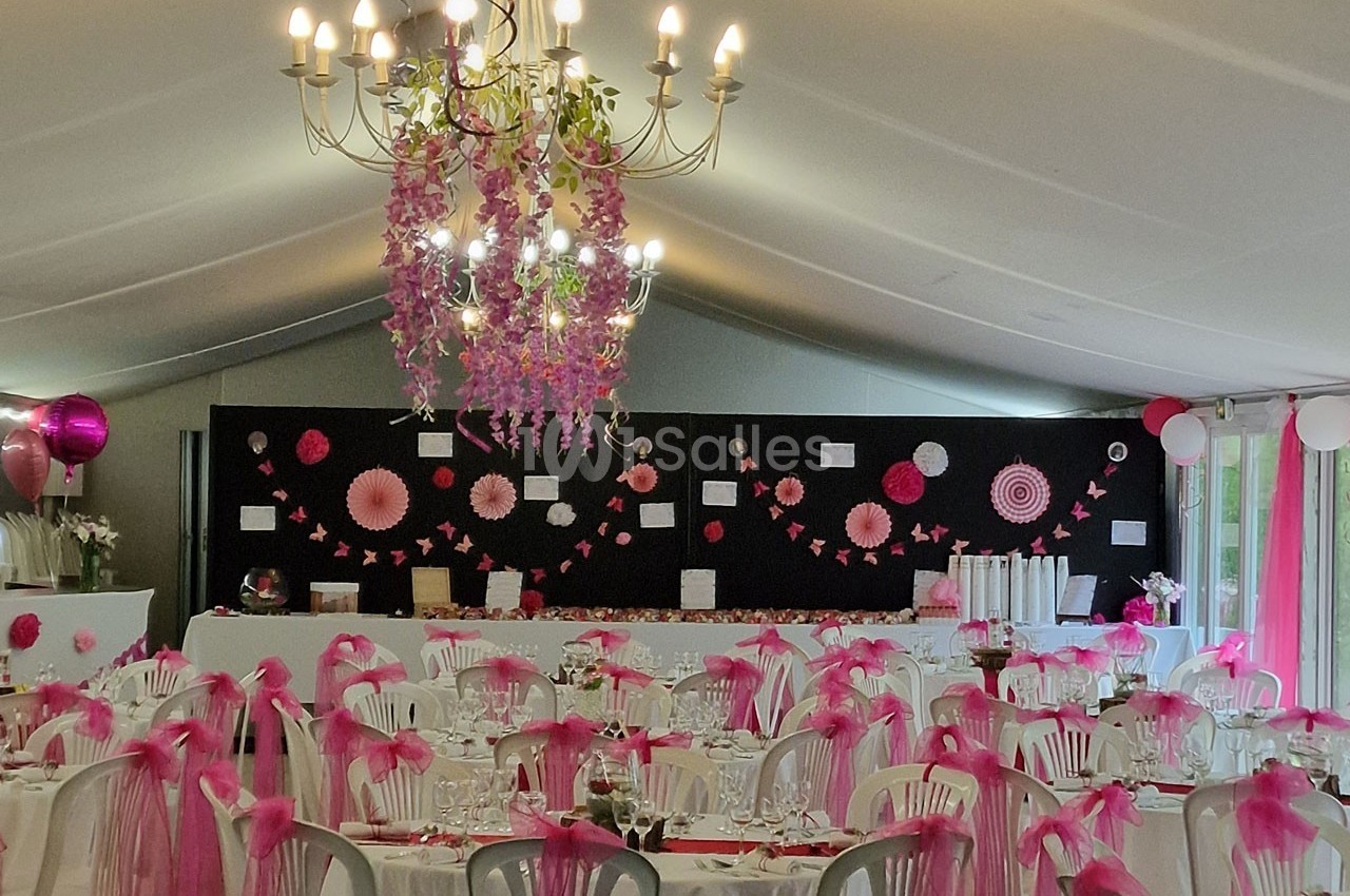Salle de réception décorée en rose et blanc avec des guirlandes, des fleurs suspendues et des tables dressées.