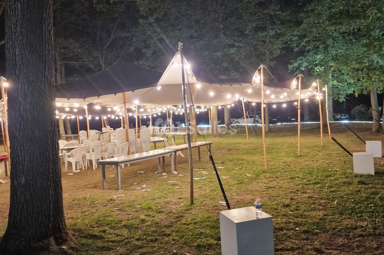 Tente éclairée par des guirlandes lumineuses dans un espace extérieur, avec des chaises et tables en plastique disposées.