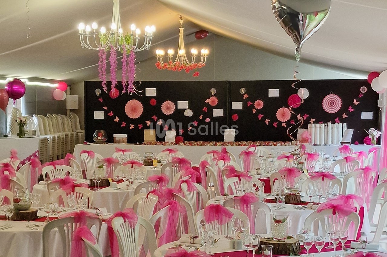 Salle de réception décorée avec des tables dressées, des chaises ornées de nœuds roses et des guirlandes suspendues.