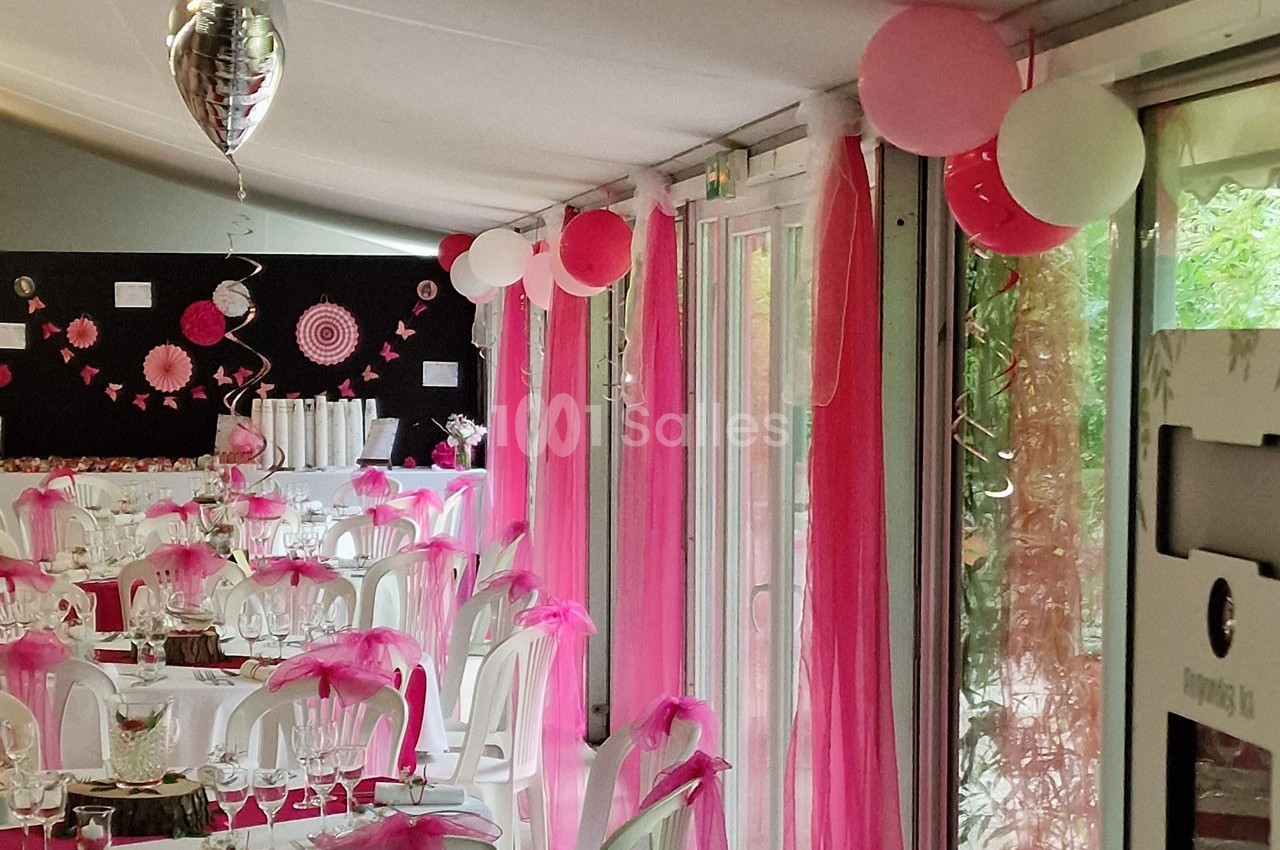 Salle décorée pour une réception avec nappes blanches, chaises ornées de rubans roses et ballons suspendus.