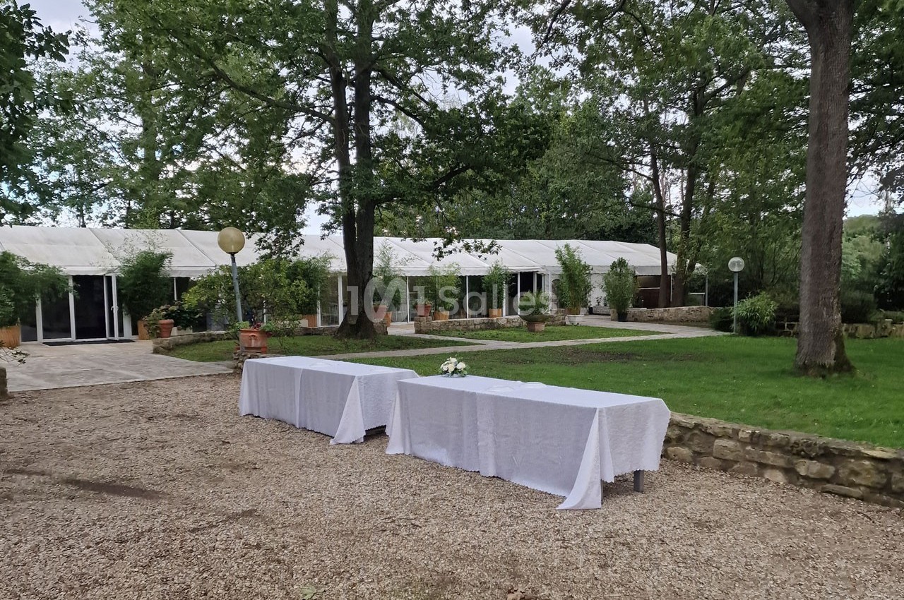 Deux tables recouvertes de nappes blanches sont disposées en extérieur, près d'une pelouse et de grandes tentes blanches.