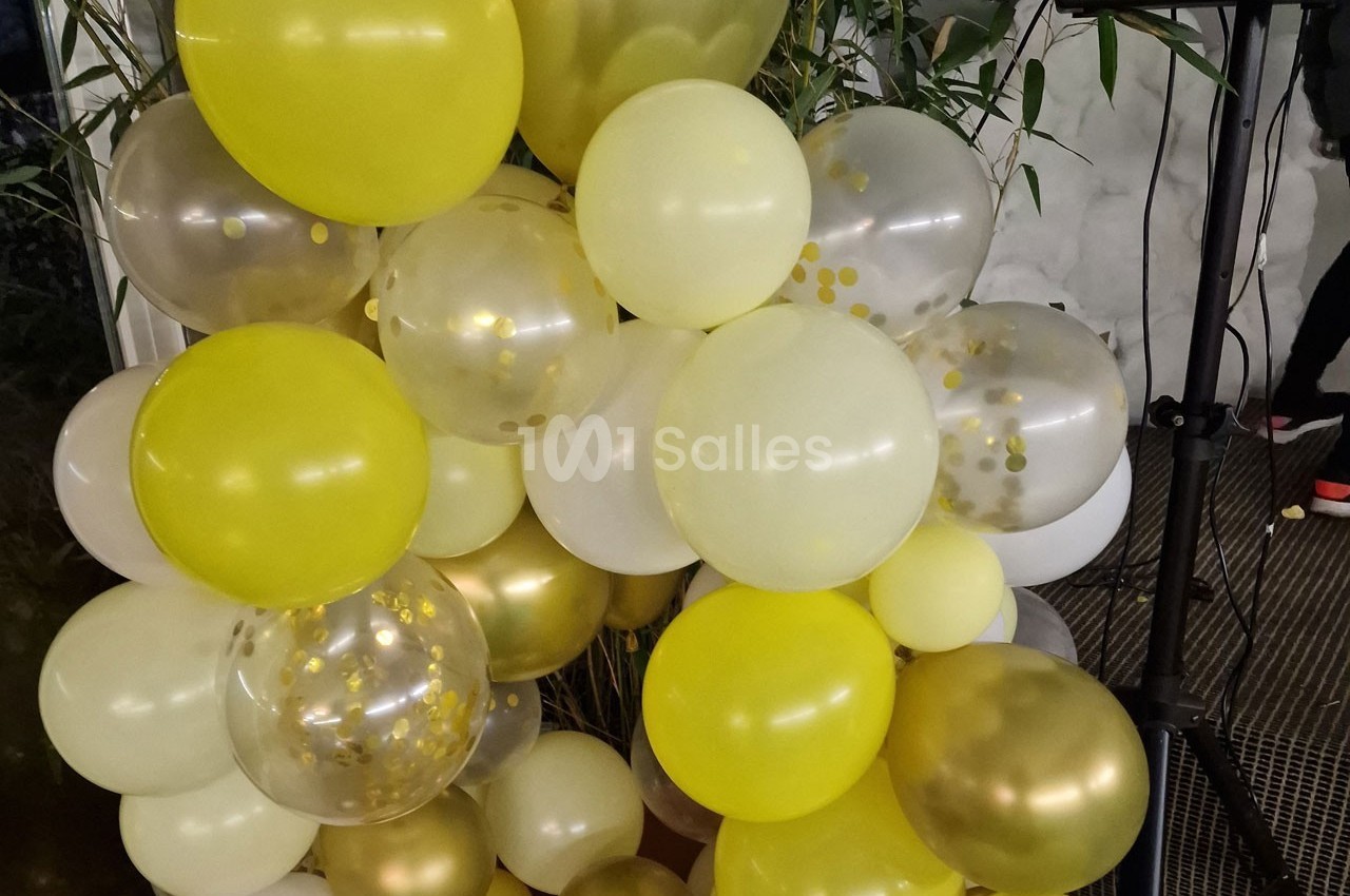 Bouquet de ballons jaunes, blancs et dorés avec des motifs de confettis, disposé devant des plantes vertes.