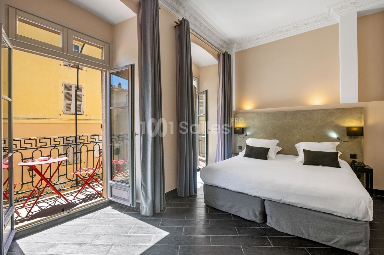 Chambre lumineuse avec lit double, rideaux gris, sol en carrelage foncé et balcon avec table et chaises rouges.