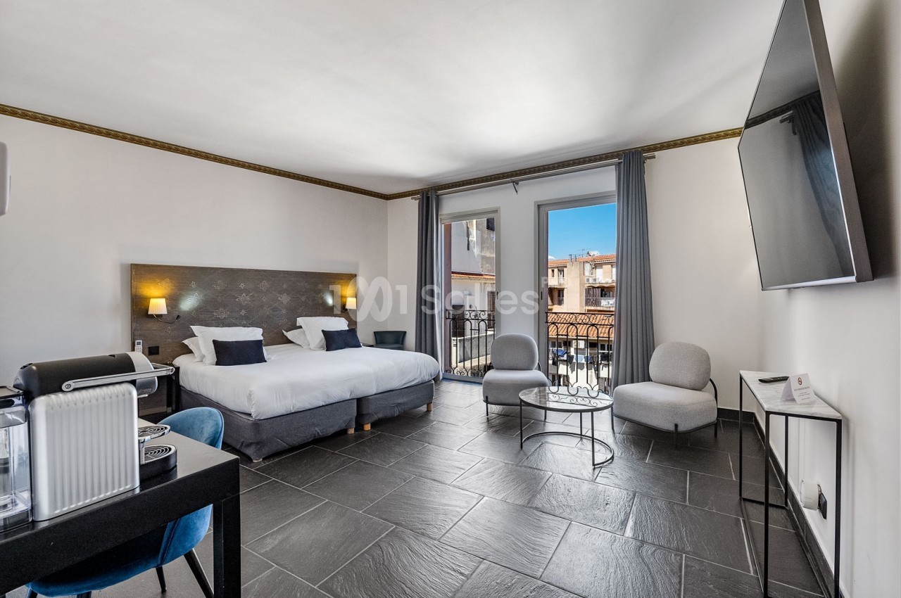 Chambre lumineuse avec lit double, coin salon, bureau, balcon et vue sur des bâtiments voisins sous un ciel bleu.