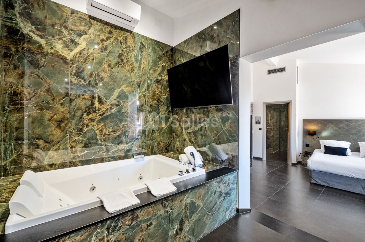 Salle de bain moderne avec baignoire à remous encastrée dans un revêtement en marbre vert, vue sur une chambre adjacente.