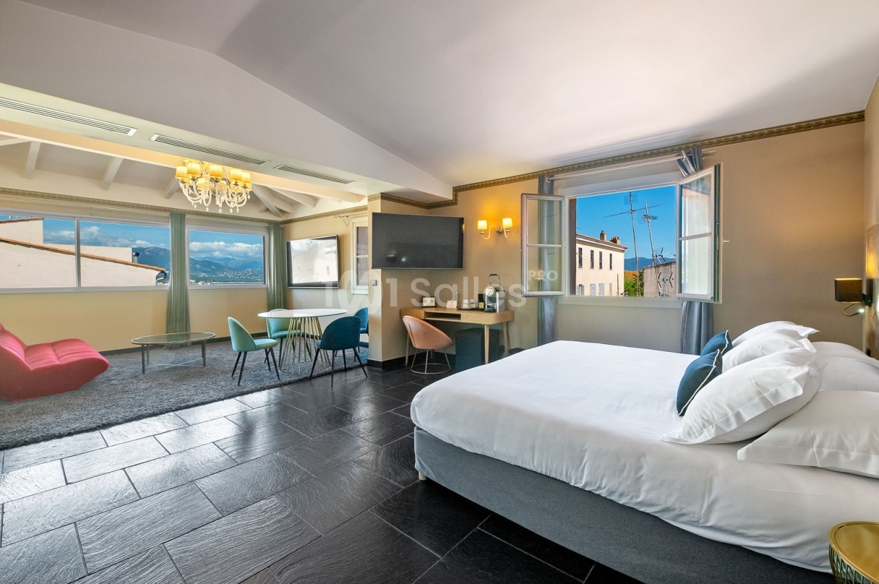 Chambre spacieuse avec lit double, coin salon, bureau, grandes fenêtres ouvertes et vue sur des bâtiments et collines.