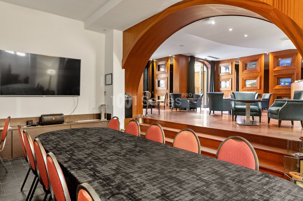Salle avec une grande table recouverte d'une nappe noire, des chaises rouges et un espace lounge en arrière-plan.